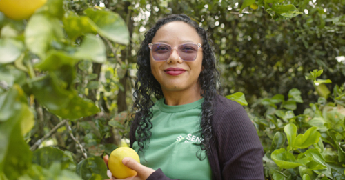 Mulher negra sorrindo enquanto segura uma laranja em uma plantação